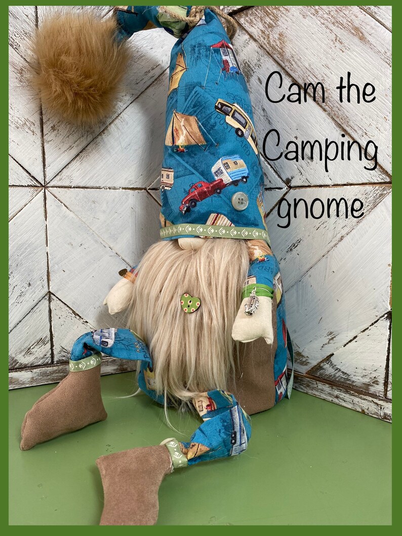May include: A blue gnome with a white beard and a hat with a camping theme. The hat has a pattern of tents, campers, and cars. The gnome is sitting on a green surface. The text "Cam the Camping Gnome" is written in white on a wooden background.