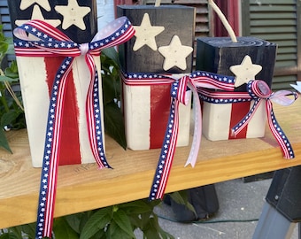 May include: Three rectangular wooden blocks painted with red, white, and blue stripes and stars. Each block features a dark blue top section with cream-colored wooden stars, a white and red striped base, a jute rope fuse, and a patriotic ribbon bow.