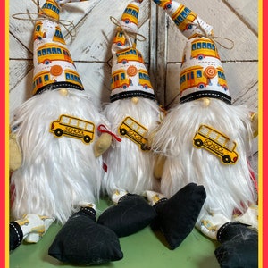 May include: Three white fuzzy gnomes with yellow school bus patterned hats and yellow school bus patches on their shirts. The gnomes are sitting on a green surface.