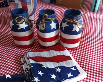 May include: Three glass jars painted with the American flag design. The jars are sitting on a red and white checkered tablecloth. There are also red, white, and blue napkins in the image.