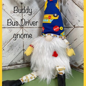 May include: A white gnome with a blue hat with yellow school buses and stop signs printed on it. The gnome is wearing a red heart and has a white beard. The gnome is sitting on a green surface with the text "Buddy Bus Driver gnome" written on a white wooden background.
