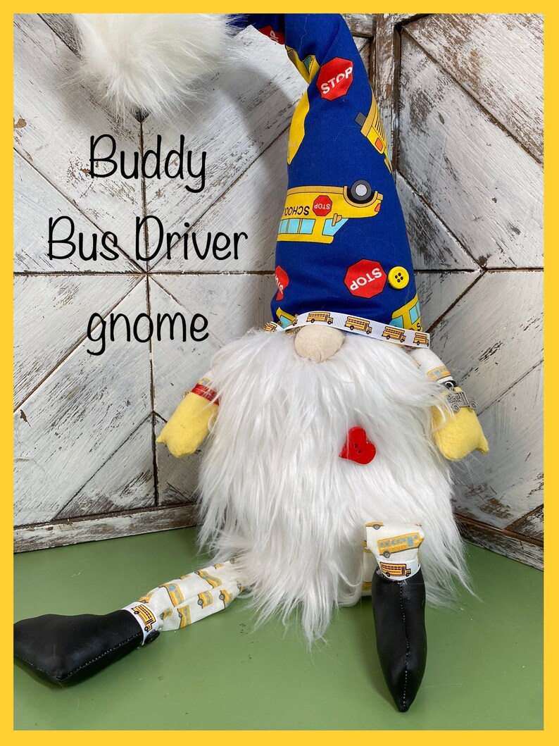 May include: A white gnome with a blue hat with yellow school buses and stop signs printed on it. The gnome is wearing a red heart and has a white beard. The gnome is sitting on a green surface with the text "Buddy Bus Driver gnome" written on a white wooden background.