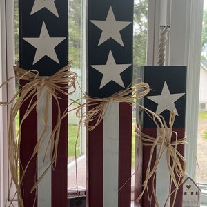 May include: Three wooden blocks decorated with red, white, and blue stripes and white stars. The blocks are tied with brown raffia ribbon and have a rope attached to the top. The blocks are arranged in descending order of size. The text "Welcome to our home" is visible on a small white house.