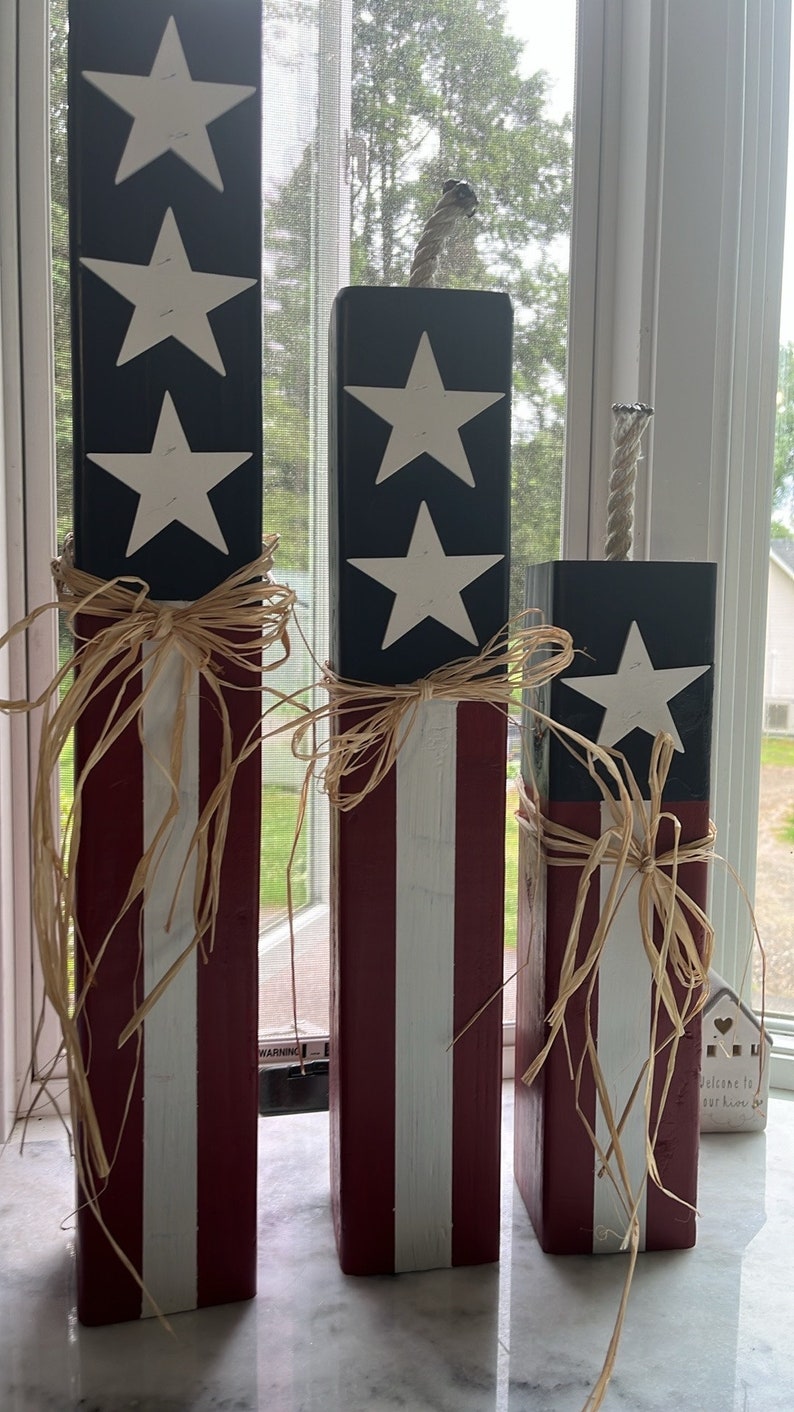 May include: Three wooden blocks decorated with red, white, and blue stripes and white stars. The blocks are tied with brown raffia ribbon and have a rope attached to the top. The blocks are arranged in descending order of size. The text "Welcome to our home" is visible on a small white house.