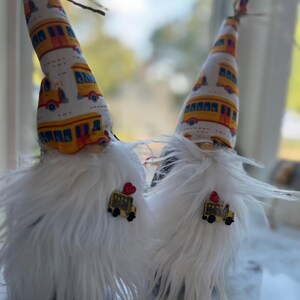 May include: Two gnomes with white fur and yellow and white hats with a school bus pattern. Each gnome has a small yellow school bus charm attached to their hat.