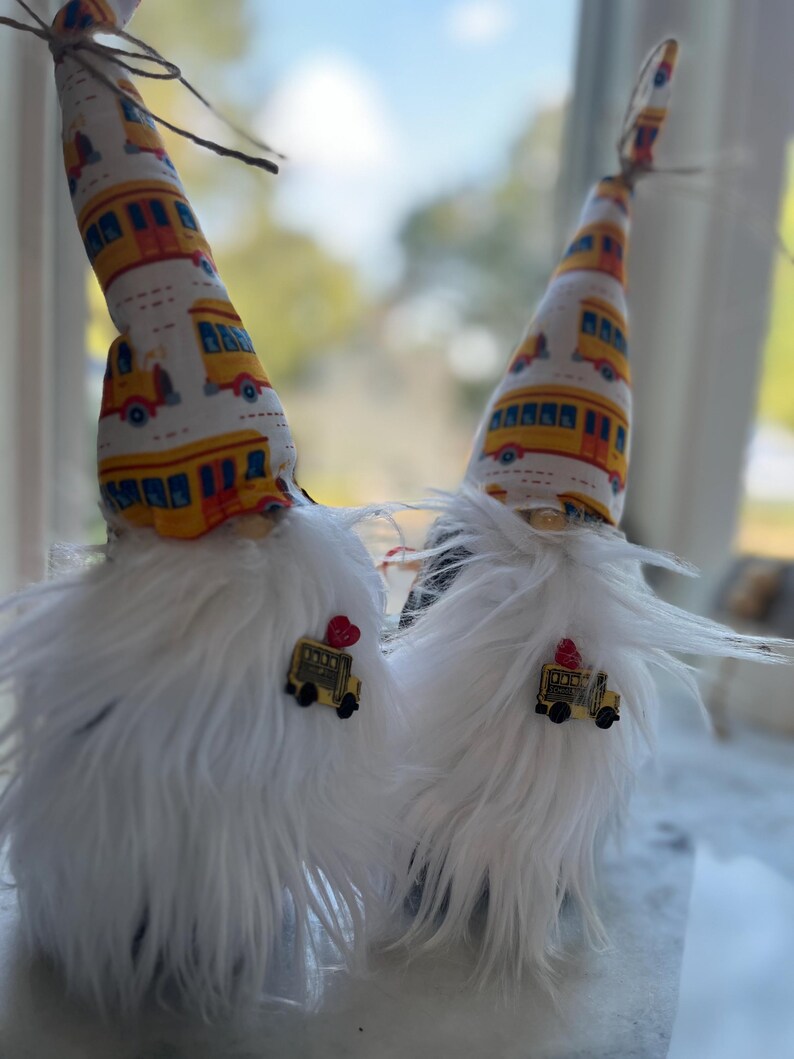 May include: Two gnomes with white fur and yellow and white hats with a school bus pattern. Each gnome has a small yellow school bus charm attached to their hat.