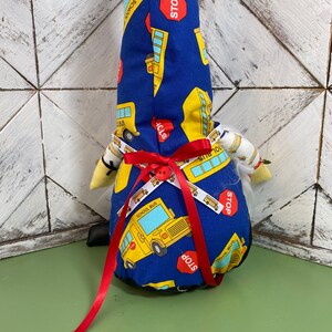 May include: A blue gnome with a white beard and a red ribbon around its waist. The gnome is wearing a hat with a school bus pattern and the words "School Bus" and "Stop" printed on it.