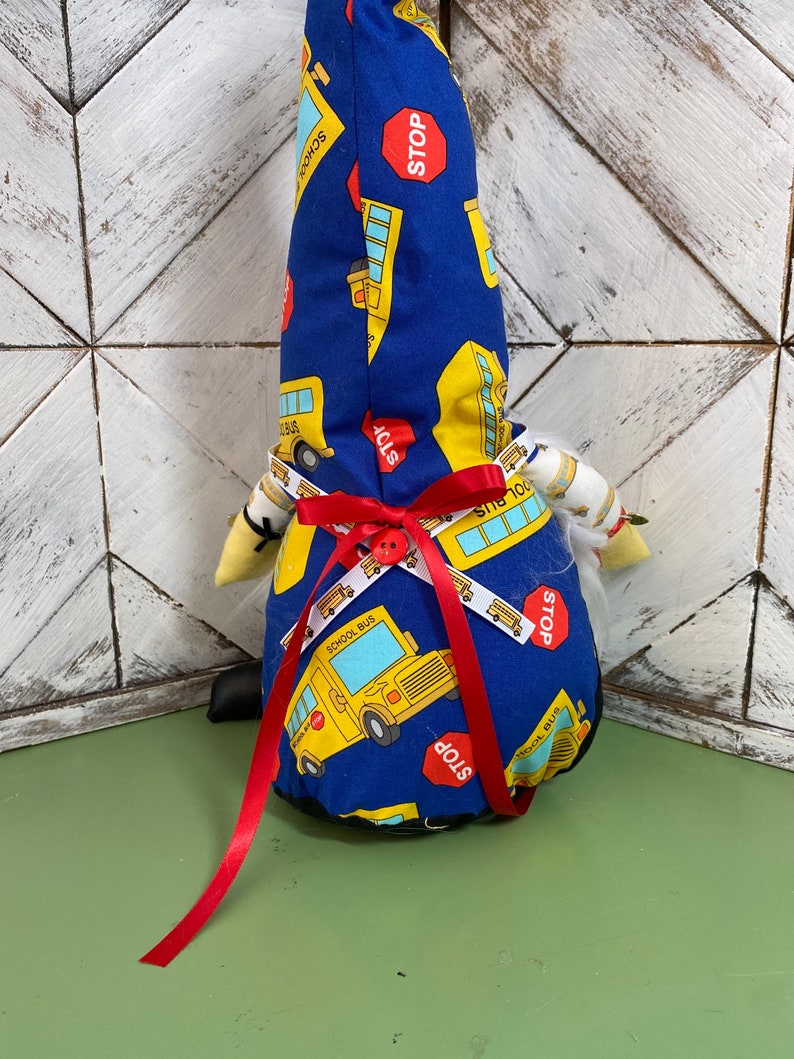 May include: A blue gnome with a white beard and a red ribbon around its waist. The gnome is wearing a hat with a school bus pattern and the words "School Bus" and "Stop" printed on it.