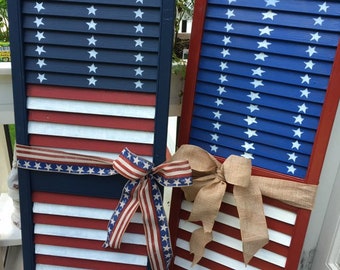 May include: Two wooden shutters painted in red, white, and blue to resemble the American flag. Each shutter has a bow tied around it. The bow on the left shutter is red, white, and blue, while the bow on the right shutter is brown.