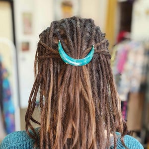 May include: A person with long brown dreadlocks wearing a teal blue sweater. A turquoise crescent moon hair clip is holding the dreadlocks in place.