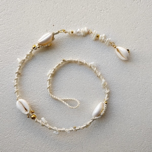 May include: A handcrafted necklace featuring a cream-colored cord base adorned with white cowrie shells, small clear crystal beads, and metallic gold-toned spherical accents. The design includes a looped end and a dangling shell charm.