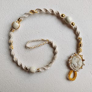 May include: A cream-colored macrame bracelet with gold accents and a circular pendant featuring a carved design. The bracelet includes white beads and a loop. The macrame is knotted in a spiral pattern.