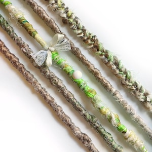 Puede incluir: Primer plano de varias rastas hechas a mano en tonos marrón, verde y crema. Algunas rastas están adornadas con tela, cintas y cuentas. Las rastas están dispuestas en diagonal sobre un fondo blanco, mostrando su textura y variaciones de color.