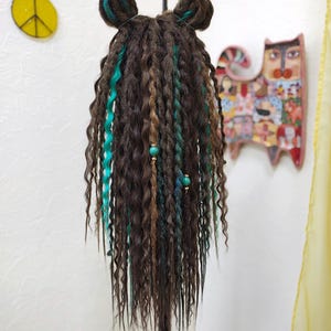May include: A brown wig with teal and brown dreadlocks. The wig is styled with two teal buns on top. The wig is on a black stand.
