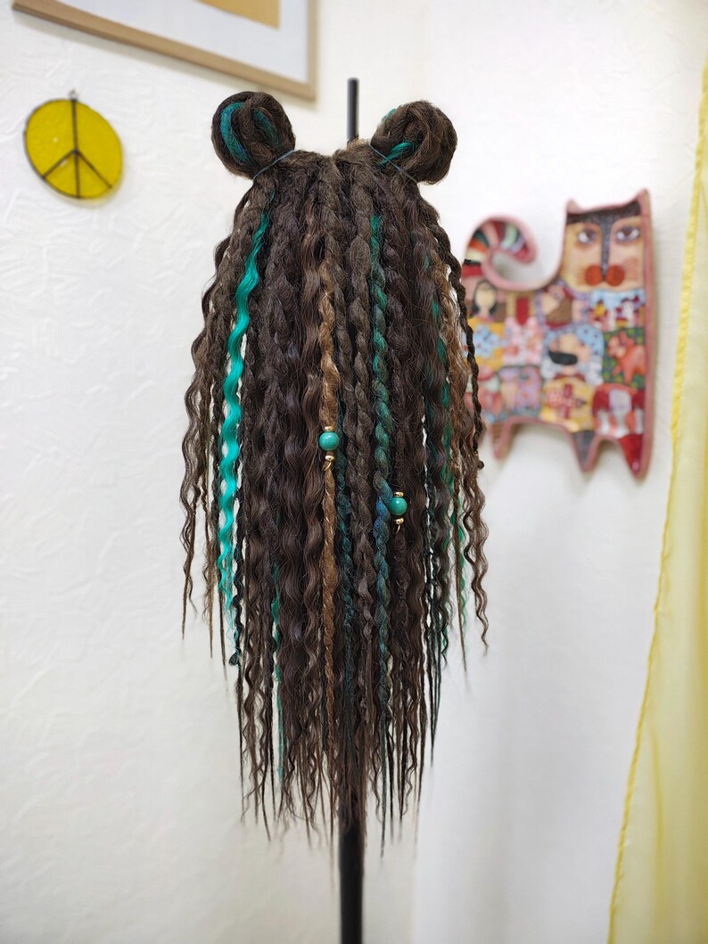 May include: A brown wig with teal and brown dreadlocks. The wig is styled with two teal buns on top. The wig is on a black stand.