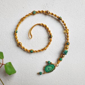 May include: A handcrafted necklace featuring a gold-colored, braided cord with green and gold beads. The necklace has a large, oval-shaped green stone pendant with gold edging. The design is intricate and detailed, with a spiral shape.