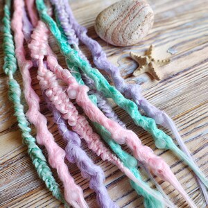 Könnte beinhalten: Pastellfarbene Dreadlocks in Rosa, Mintgrün und Lavendel. Die handgefertigten Haarverlängerungen sind auf einer Holzoberfläche angeordnet, mit einem kleinen Stein und einem Seestern als Akzent. Die Dreads sind ein einzigartiges Haaraccessoire.