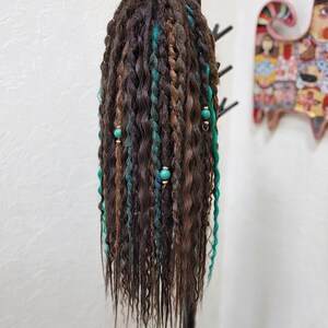 May include: A long, brown wig with teal and green dreadlocks. The wig is styled with beads and is hanging on a black stand.