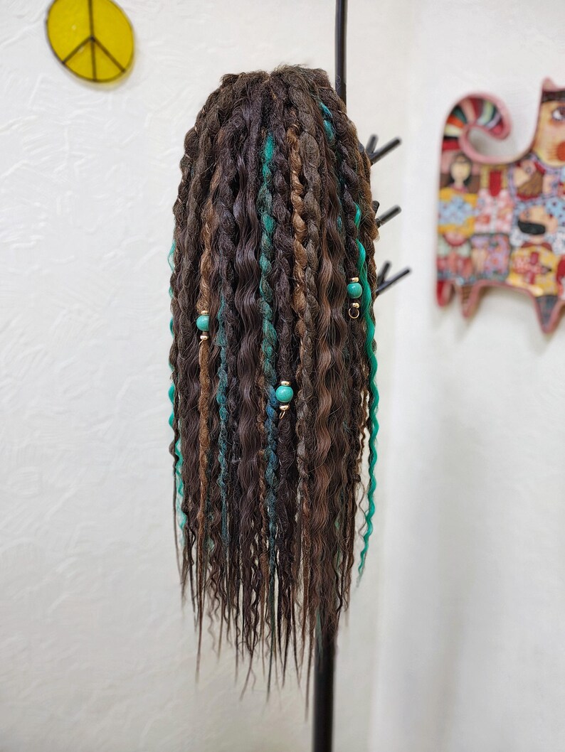 May include: A long, brown wig with teal and green dreadlocks. The wig is styled with beads and is hanging on a black stand.