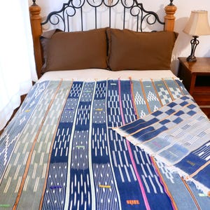 May include: A woven textile with a geometric pattern in shades of blue, white, and pink is draped over a bed. The bed has brown pillows and a decorative metal headboard. A matching piece of the textile is folded on the bed.