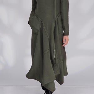 Può includere: Un cappotto verde oliva con cappuccio, cerniera a tutta lunghezza e ampie tasche. Il cappotto ha un design asimmetrico unico e una gonna svasata. Il cappuccio è foderato con un tessuto più scuro. Il cappotto è realizzato in un materiale spesso e caldo.