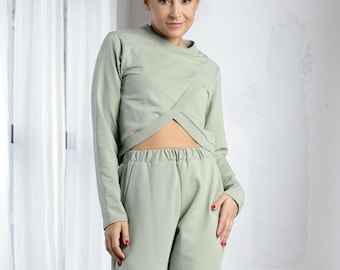 Puede incluir: Un conjunto de dos piezas de color verde salvia con una blusa de manga larga y corta y pantalones a juego. La blusa tiene un diseño cruzado en la parte delantera.