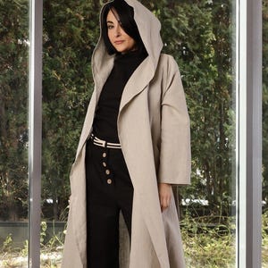 Könnte beinhalten: Ein langer, beigefarbener Mantel mit Kapuze und fließendem Design. Der Mantel wird über einem schwarzen Rollkragenpullover, einer schwarzen Hose und einem weißen Gürtel getragen. Die Hose hat große Knöpfe und ein weites Bein. Das Outfit befindet sich vor einem großen Fenster und Grün.