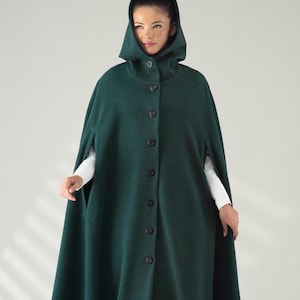 Könnte beinhalten: Ein langer, dunkelgrüner Kapuzenmantel mit Knopfleiste. Der Umhang ist bodenlang und hat ein weites, fließendes Design. Das Model trägt darunter ein weißes Langarmshirt. Der Umhang ist aus einem dicken, warmen Material gefertigt.