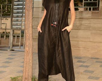 May include: A woman wearing a black linen jumpsuit with a long black beaded necklace and white sneakers. She is standing in front of a tree and a fence.