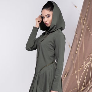 Puede incluir: Una chaqueta larga con capucha de color verde oliva, con cierre de cremallera y un detalle de cremallera diagonal. La chaqueta tiene un corte holgado y un dobladillo ligeramente asimétrico. La modelo lleva pantalones blancos y zapatos plateados.