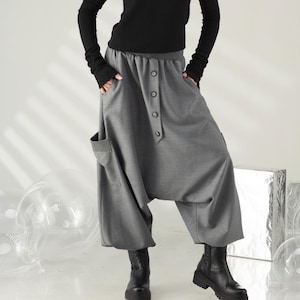 May include: A woman wearing a black long-sleeved turtleneck top and gray harem pants with large pockets and button details. She is also wearing black boots. The background is a light gray.