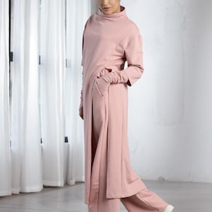 Pode incluir: Um macacão comprido, rosa, solto, com gola alta e mangas compridas. O macacão tem um caimento relaxado e uma silhueta fluida. A modelo está usando tênis brancos.