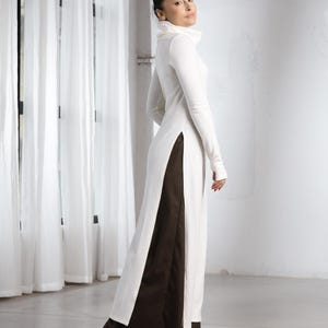May include: A long, white dress with a high collar and long sleeves. The dress features a dark brown panel on the sides and is paired with black heels. The outfit is a modern take on a classic design.