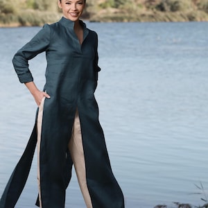 Puede incluir: Una túnica de color verde azulado con cuello alto y mangas largas, combinada con pantalones beige de pernera ancha. El vestido tiene una abertura frontal y se lleva al aire libre cerca de un cuerpo de agua.