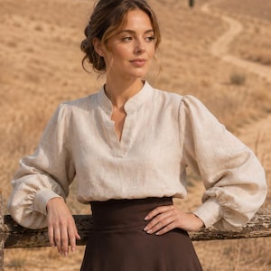 May include: A beige long-sleeved blouse with a mandarin collar and a V-neckline, paired with a high-waisted dark brown maxi skirt. The blouse features voluminous sleeves with fitted cuffs, while the skirt has a smooth, flared silhouette.