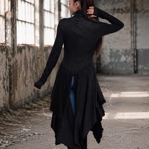 May include: A long-sleeved black tunic with a high turtleneck collar and thumbhole cuffs. The garment features a fitted bodice that transitions into a flared, asymmetrical hemline with layered fabric panels, paired with blue denim jeans and black boots.