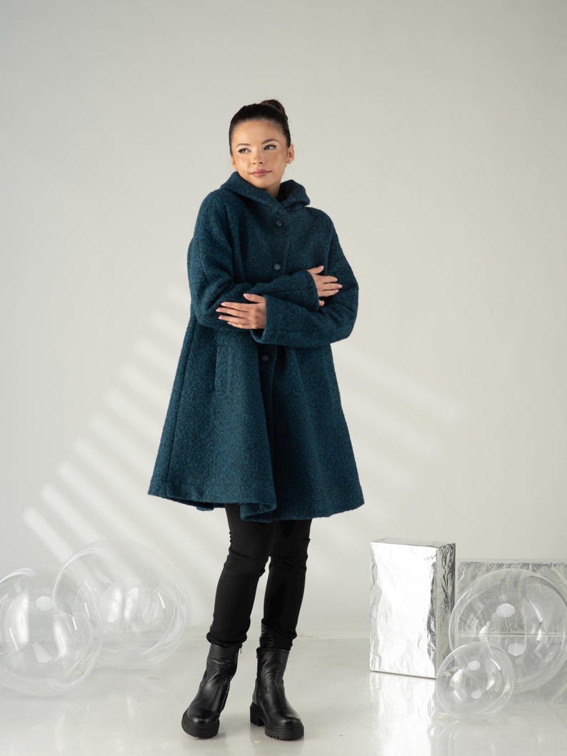 May include: A teal hooded coat with a button-up front. The coat has a flared silhouette and is paired with black pants and black boots. The coat is made of a textured fabric.