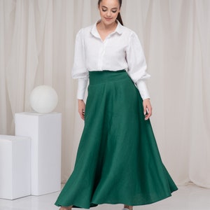 May include: A woman wearing a white button-down shirt with long sleeves and a green maxi skirt. She is standing on a white surface with white curtains in the background.