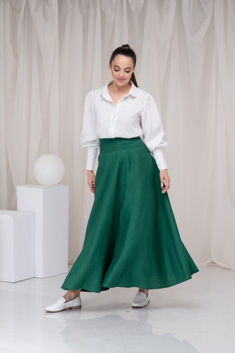 May include: A woman wearing a white button-down shirt with long sleeves and a green maxi skirt. She is standing on a white surface with white curtains in the background.