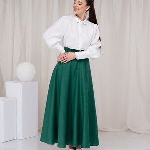 May include: A woman wearing a white collared shirt with puffed sleeves and a long, dark green skirt. She is standing in front of a white backdrop.