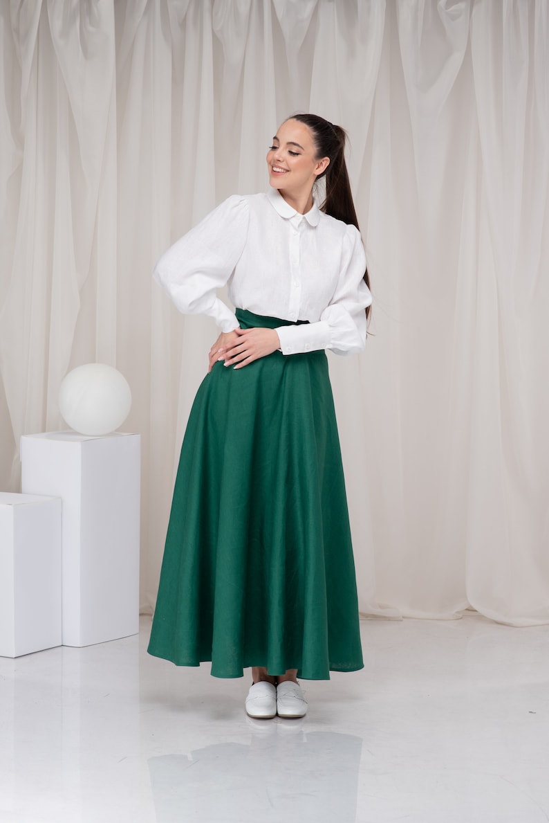 May include: A woman wearing a white collared shirt with puffed sleeves and a long, dark green skirt. She is standing in front of a white backdrop.