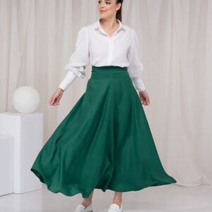 May include: A woman wearing a white button-down shirt and a long, dark green skirt. She is standing in front of a white backdrop and is looking to the right.