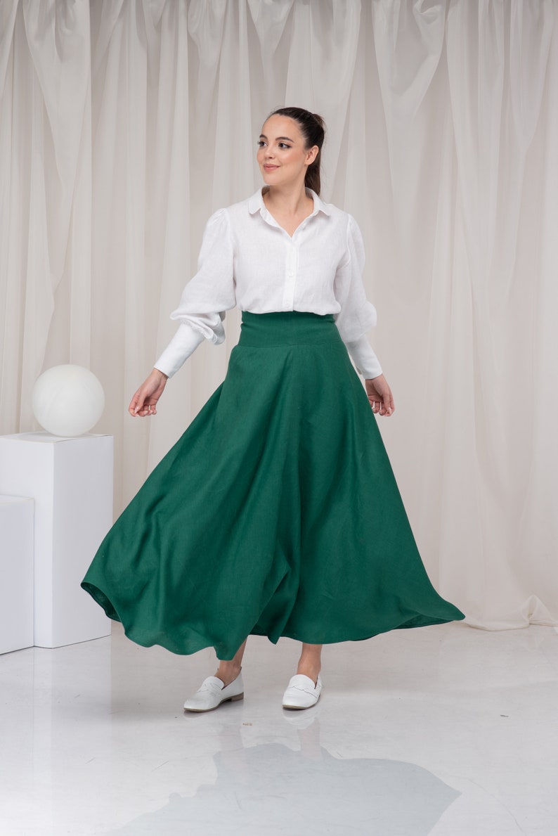 May include: A woman wearing a white button-down shirt and a long, dark green skirt. She is standing in front of a white backdrop and is looking to the right.