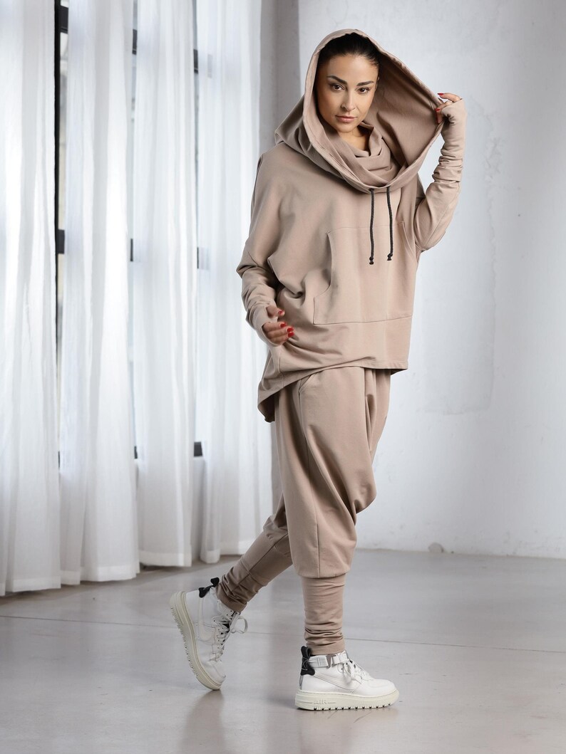 K&ouml;nnte beinhalten: Ein beiger Kapuzenpullover mit Kordelzug und einer K&auml;ngurutasche. Der Pullover wird mit einer passenden beigen Hose kombiniert. Das Model tr&auml;gt wei&szlig;e Sneaker.