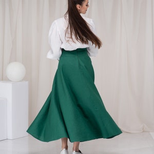 May include: A woman wearing a white blouse and a long, green skirt. She is standing in front of a white backdrop.