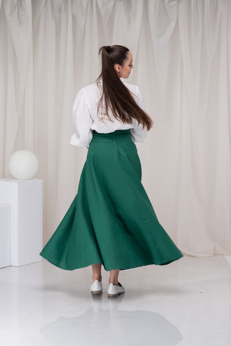 May include: A woman wearing a white blouse and a long, green skirt. She is standing in front of a white backdrop.