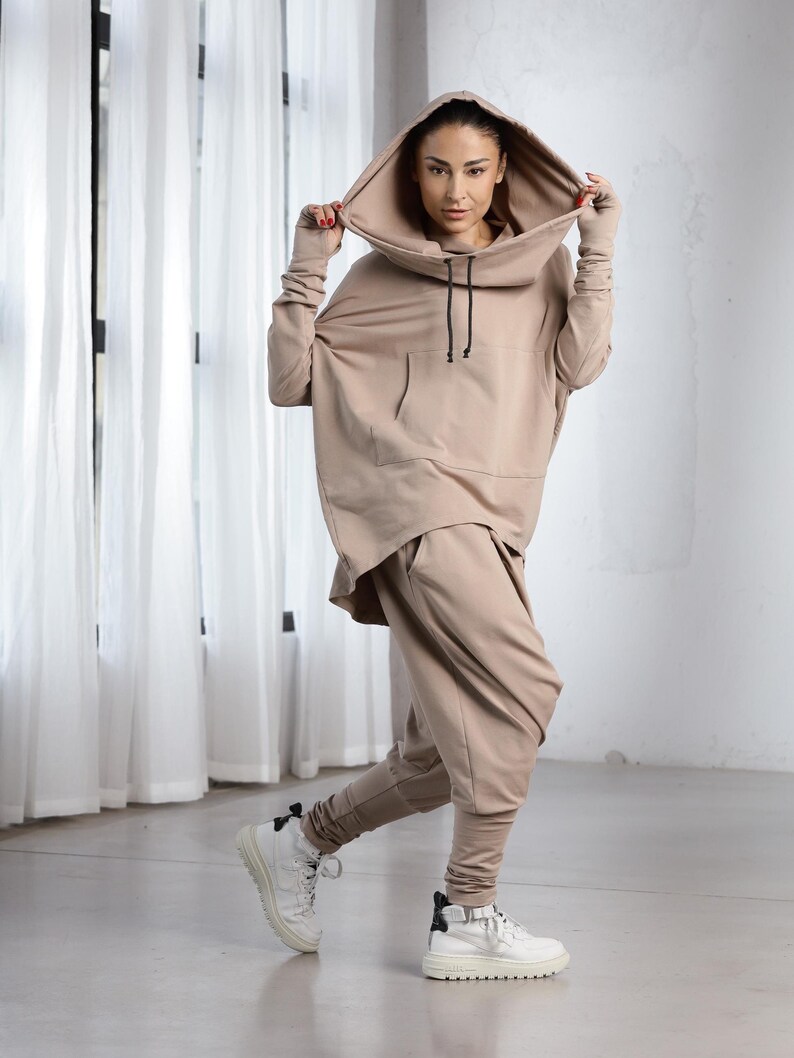 K&ouml;nnte beinhalten: Ein beiger Kapuzenpullover und eine passende Hose mit Kordelzug. Der Pullover hat eine gro&szlig;e Kapuze und eine K&auml;ngurutasche. Die Hose hat eine lockere Passform.