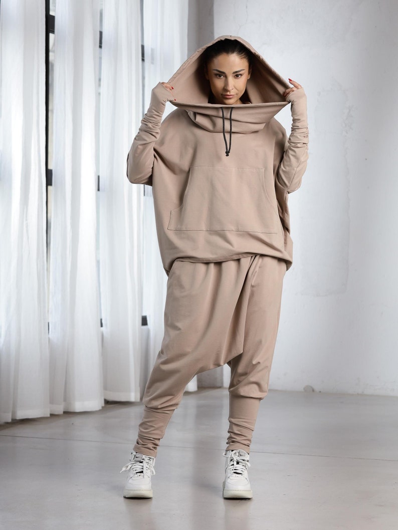 K&ouml;nnte beinhalten: Ein beiger Kapuzenpullover mit hohem Kragen und einem Kordelzug. Der Pullover hat eine gro&szlig;e Tasche auf der Vorderseite und lange &Auml;rmel. Das Model tr&auml;gt dazu eine passende beige Hose mit lockerer Passform.