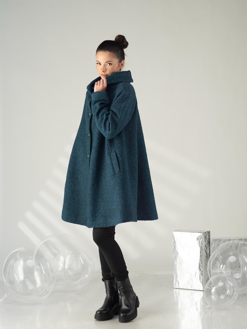 May include: A teal-colored, oversized coat with a high collar and button closure. The coat has a flared silhouette and is paired with black leggings and boots. The coat's texture appears to be a soft, woven fabric, suitable for cooler weather.