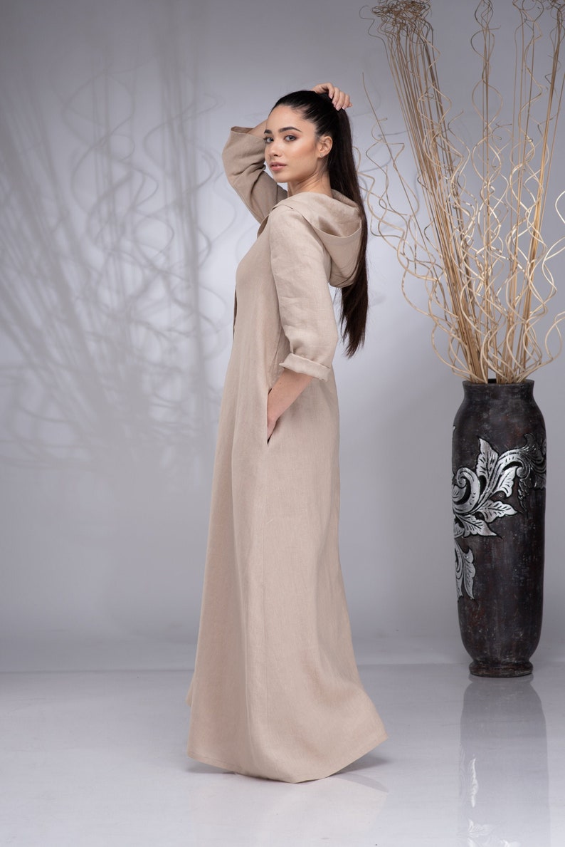 May include: A beige linen maxi dress with a hood and long sleeves. The dress has a relaxed fit and a flowing silhouette.